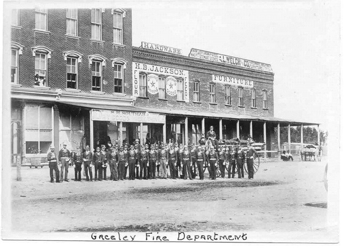 A vintage black and white photo showcasing the Greeley Fire Department with members standing in front of historic downtown buildings.