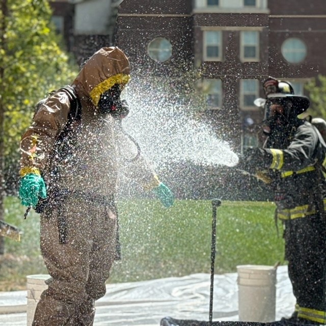 A Greeley firefighter in full bunker gear and helmet sprays water from a hose at another Greeley firefighter who is wearing a Hazmat yellow and tan suit with green gloves and a gas mask. White buckets sit on the ground, and the background features grass, trees, and a brick building with windows.