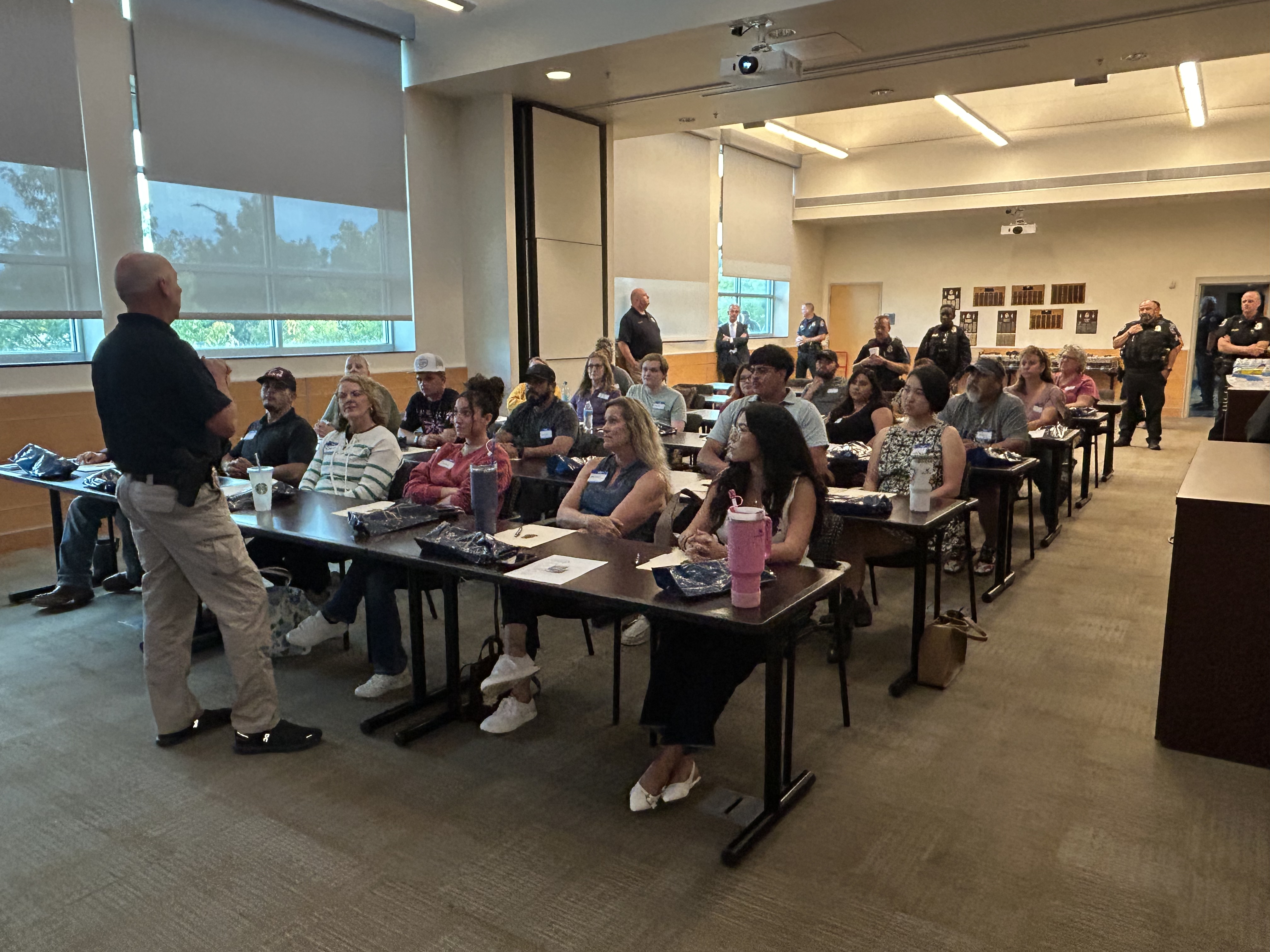 Greeley Police Chief Adam Turk speaks to members of the Greeley community during the Citizen Police Academy. There are six rows of tables with six people per row. Police officers are standing at the back of the room. 