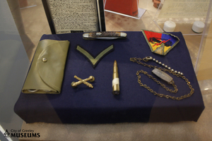 Various keepsakes, including a pocket knife, necklace, and patches displayed on a navy blue museum display.