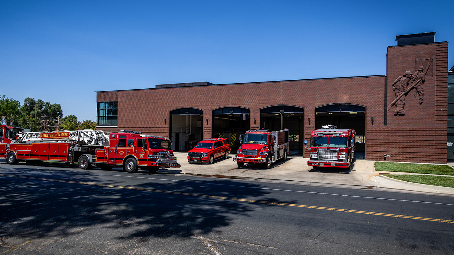 A picture taken from the front of Greeley Fire Station 1 that shows the building with 4 bays open and 4 fire trucks staged in front of the station. 