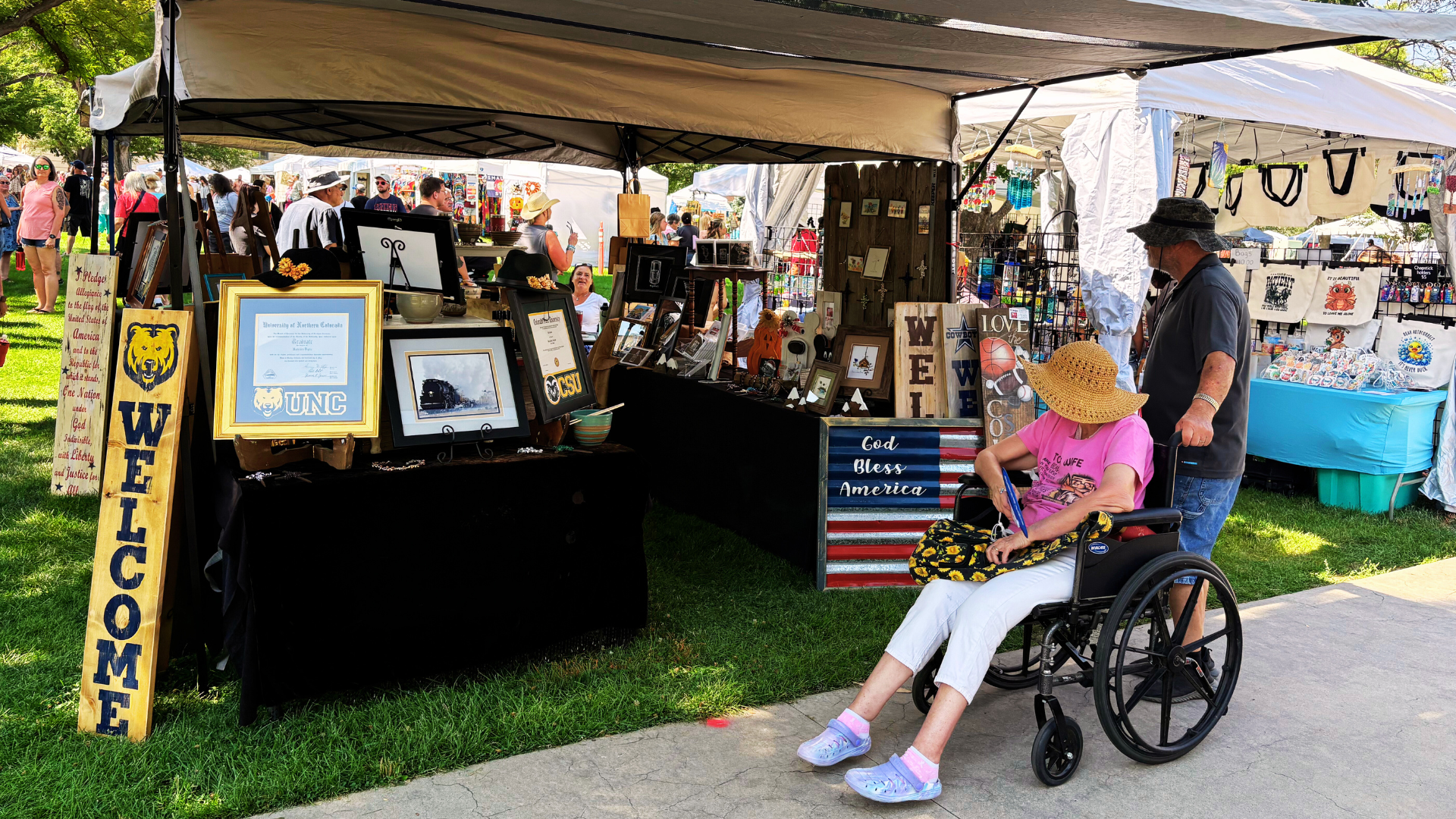 A person in a wheelchair and friend look at an arts booth with welcome signs and other signs featuring UNC logo at the Greeley Arts Picnic on a sunny day.