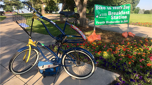 A vibrant scene showcasing a bike parked near a colorful flowerbed with signage promoting Bike to Work Day's breakfast station.