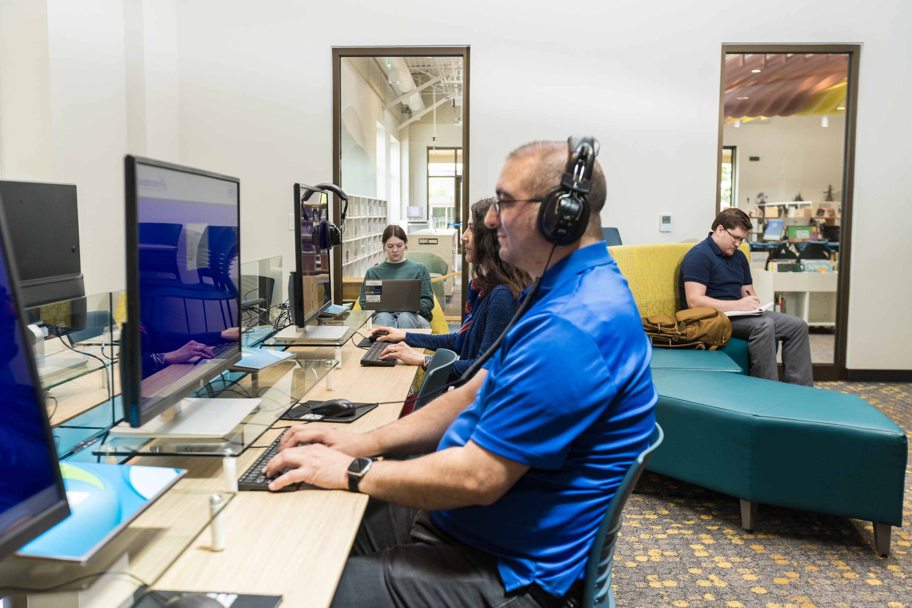 This image depicts Greeley residents using the LINC Library learning laboratory.  In the foreground of the photo, a man and woman are wearing headphones and typing on a computer at a LINC Library workstation.  Another man and woman are sitting in the background of the photo.  The man is writing in a notebook and the woman is working on a laptop computer as she sits in an oversized yellow chair.  Behind these people are windows that look into the main library space.