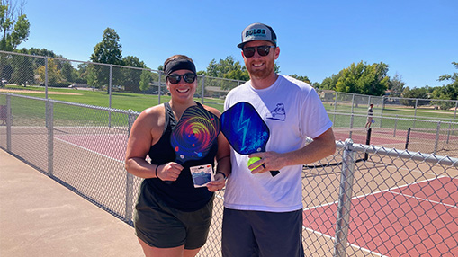 A couple holding pickleball paddles standing near a court on a sunny day, representing an engaging outdoor activity.