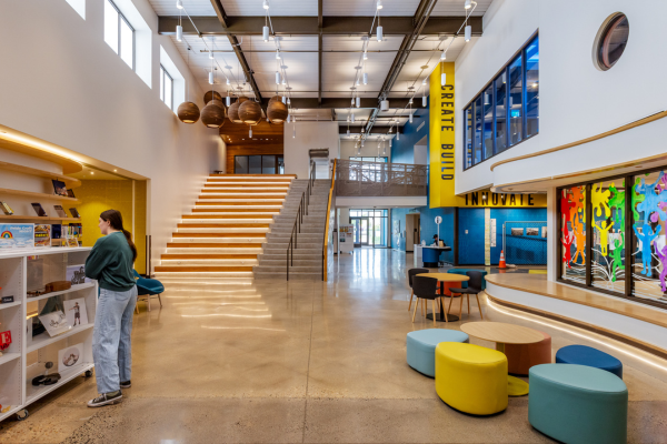 This image depicts the interior lobby of the LINC Library in Greeley, Colorado.  To the right are interior windows adorned with bright red, orange, green, blue and purple stickers in the shapes of children.  In front of those windows are brown tables with light blue, dark blue, and yellow chairs.  A woman is standing to the left of those tables, browsing decorative items on white shelves.  Behind the woman is a staircase taking visitors to the second level of the library.  Modern brown pendant lights hang directly above the staircase.