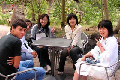 A group of individuals casually gathered outdoors around a table, surrounded by greenery.