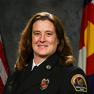 Teresa Hinz in official fire department uniform, standing against a backdrop with flags.