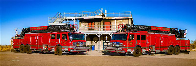 Two red firetrucks park near a training structure 
