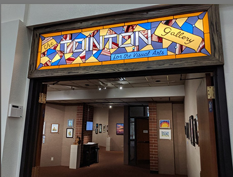 The image showcases a colorful entrance to a visual arts gallery featuring various framed artwork and sculptures inside.