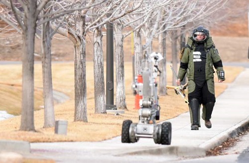 A bomb disposal technician walks near a robotic device, dressed in protective gear, during an operation in a public area.