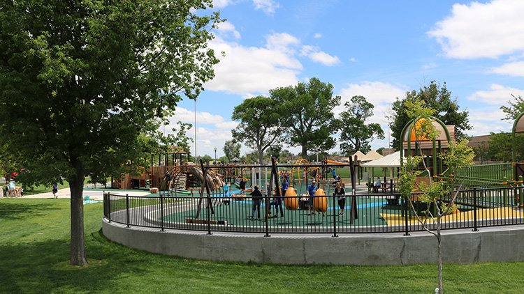 Green grass, trees, and a black metal fence surround a yellow, green, and blue playground with accessible swings and ramps, offering a vibrant recreational space.