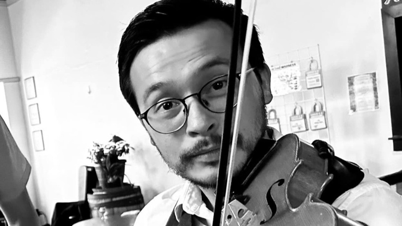 A man wearing glasses and a vest over a collared shirt plays the violin indoors. The background features tables, chairs, and wall decorations.