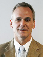 Formal image of an individual in business attire featuring a beige suit jacket and light blue tie.