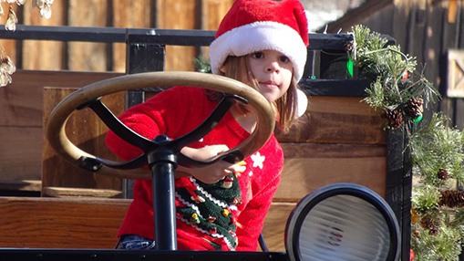 A child enjoying a holiday-themed activity dressed in a festive sweater and Santa hat, seated in a decorated vintage vehicle amid a rustic setting.
