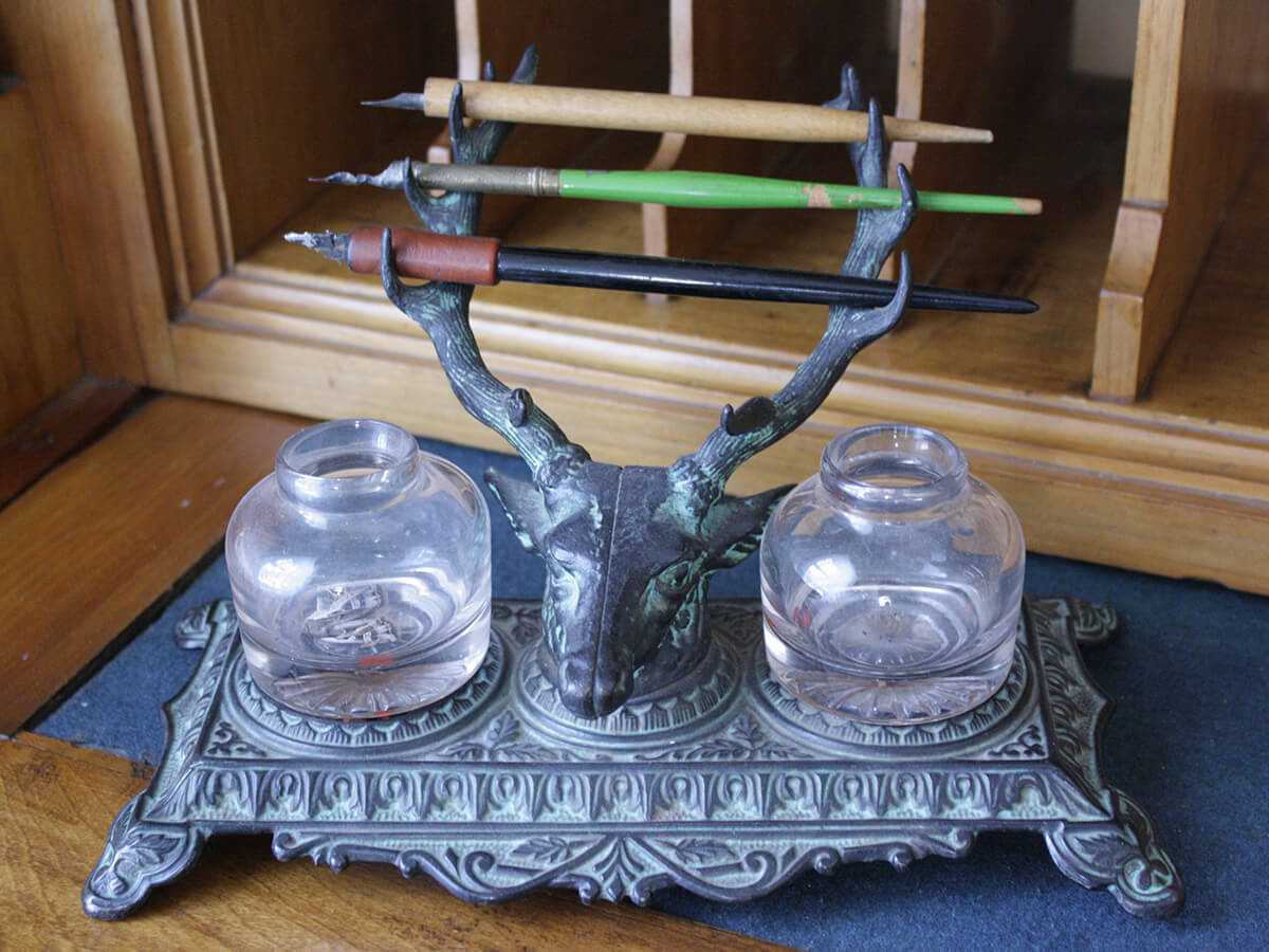 A small, decorative inkwell stand with legs featuring a stag's head, which holds three calligraphy pens in its antlers, and is flanked by two small glass jars
