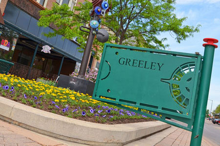 A vibrant area in Greeley with colorful flowers, a decorative sign, and nearby shops, representing community and activity.