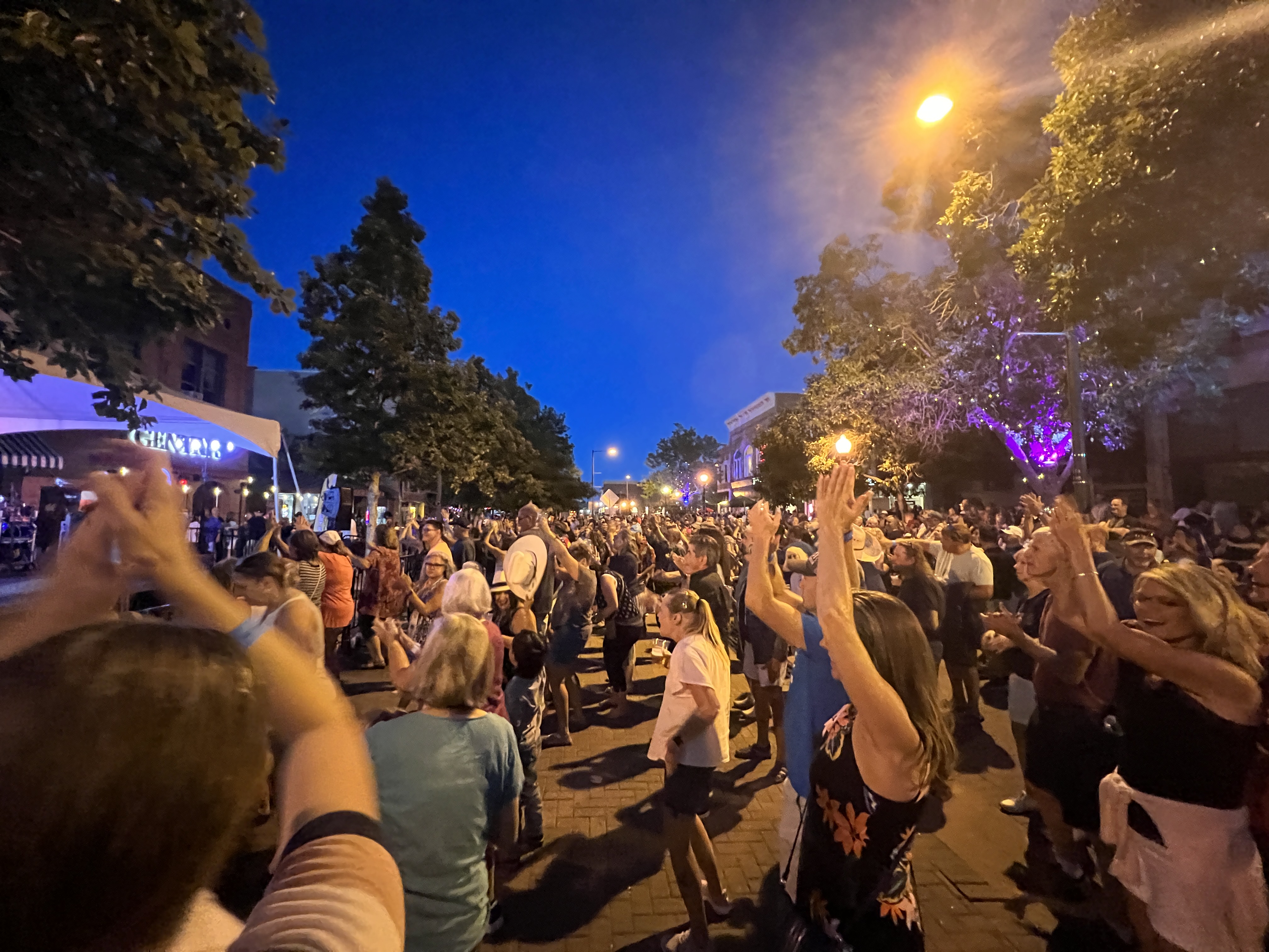 A large crowd of Greeley residents and visitors are enjoying a free, nighttime outdoor concert as part of the Greeley Friday Nights series.  A blue and white tent covers a stage area where a band plays music for hundreds of audience members who are standing, dancing, and clapping to the music.  The audience is standing in streets paved with red brick and lined with mature trees.
