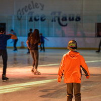 A young child in an orange shirt and backward hat skates towards others on the brightly colored Greeley Ice Haus rink.