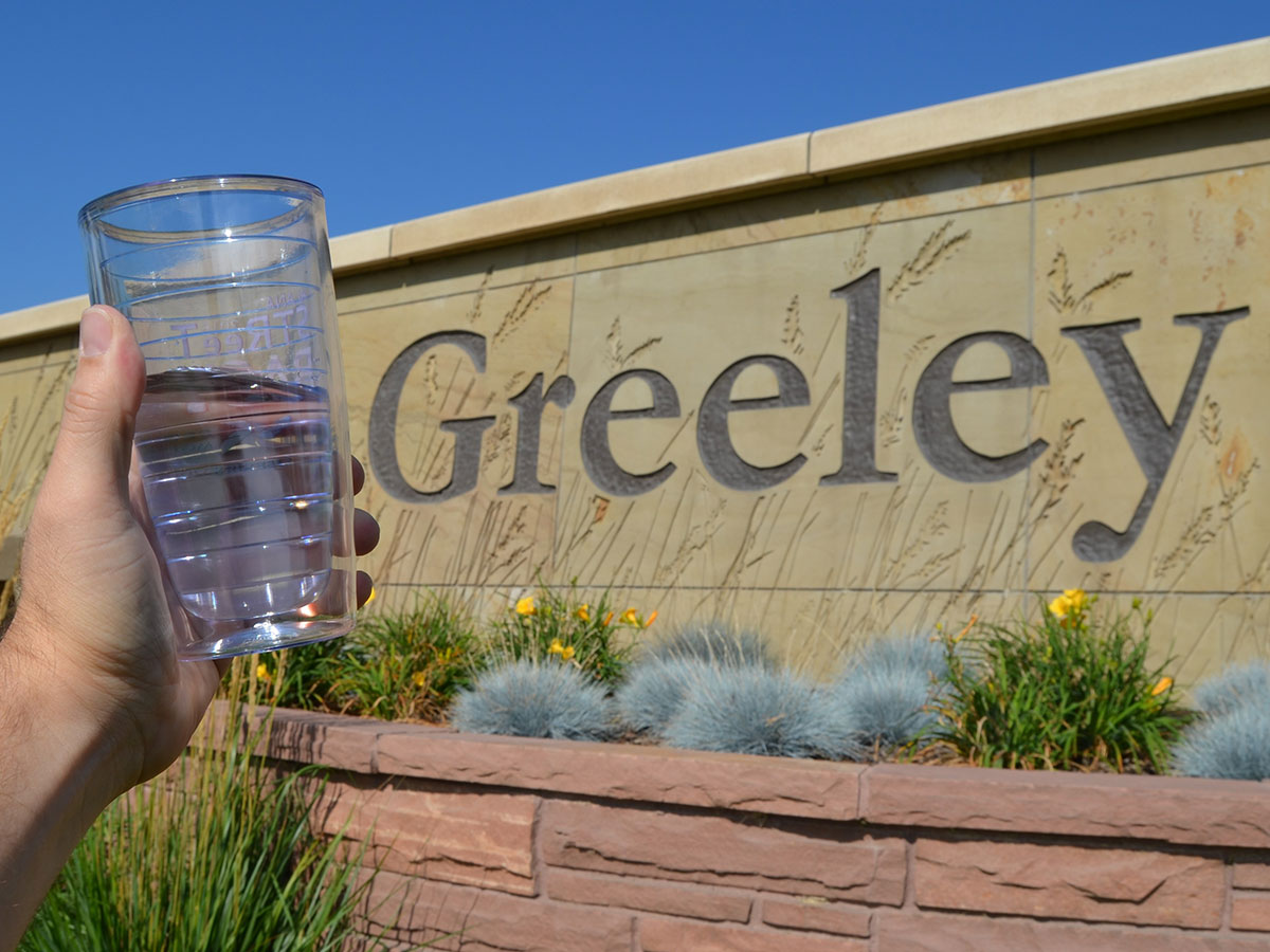 A hand holds up a large clear cup half full of water in front of an engraved Greeley city sign