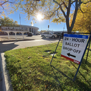 A sign outside Greeley City Hall indicating the location for 24-hour ballot drop-off, surrounded by autumn trees and parked cars.