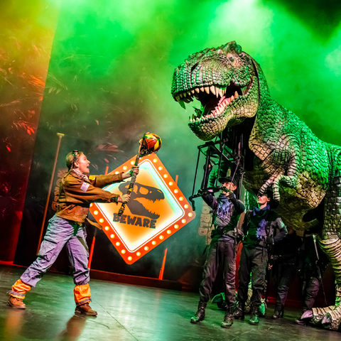 A performer tires to ward off a large T-Rex puppet operated by three other performers in front of a Beware of T Rex sign on stage in the Dinosaur World Live show