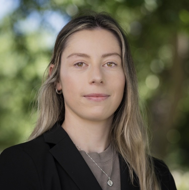 Professional headshot of Lauren Smith, Economic Development Coordinator.  The woman is wearing a black suit jacket over a tan shirt and is wearing a necklace with a round pendant containing the letter "L".  