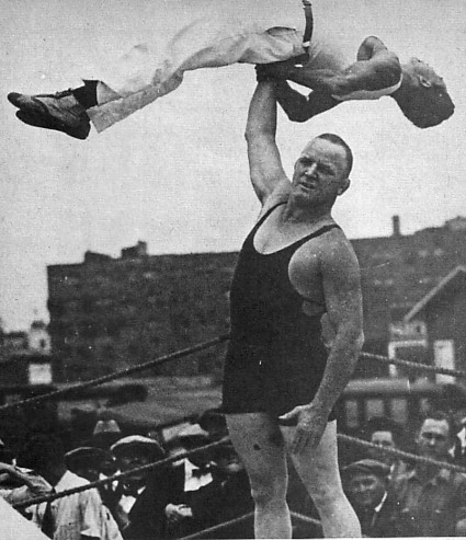 Old black and white photo of a wrestling holding another wrestler up in the air with one arm.