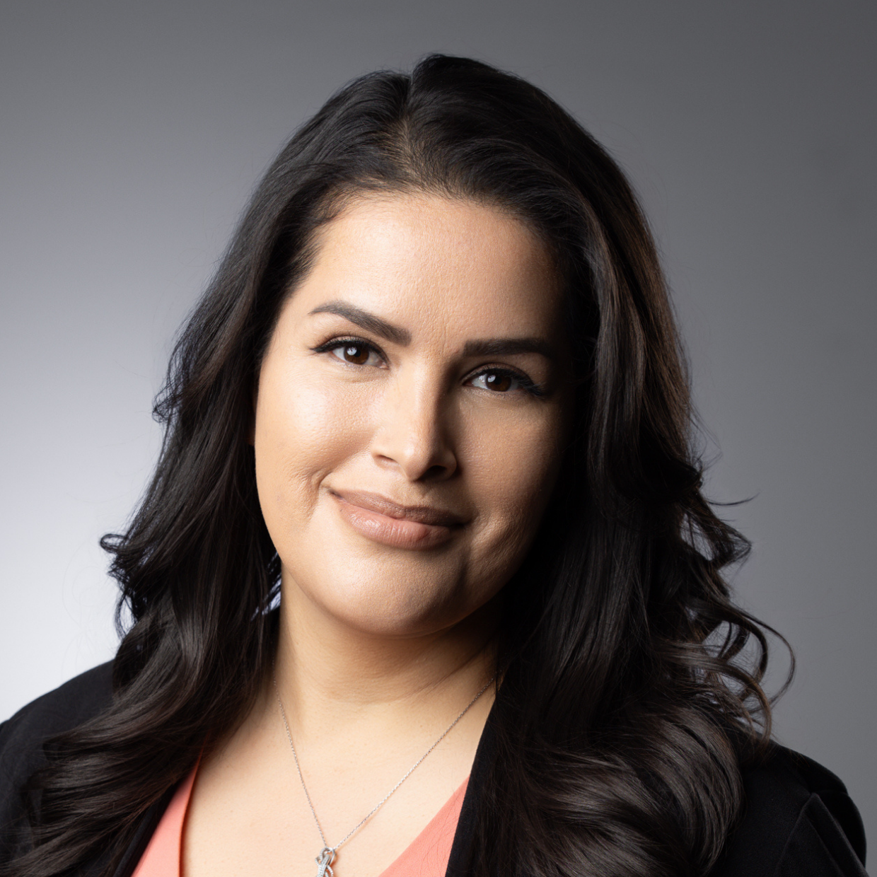 This image depicts a professional headshot of Real Estate Specialist Monique Perez.  Monique is shown in a dark suit jacket with a peach-colored v-neck blouse beneath.