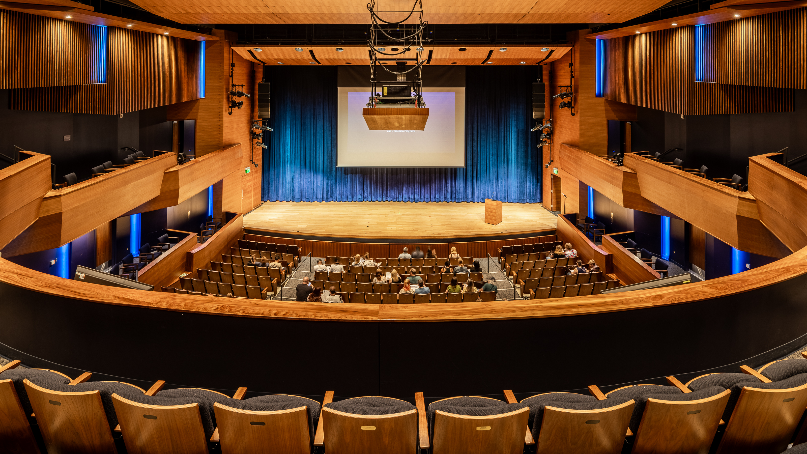 Interior view of a University of Northern Colorado performance hall with wood balconies, blue stage curtains, theater seating, and a projection screen.