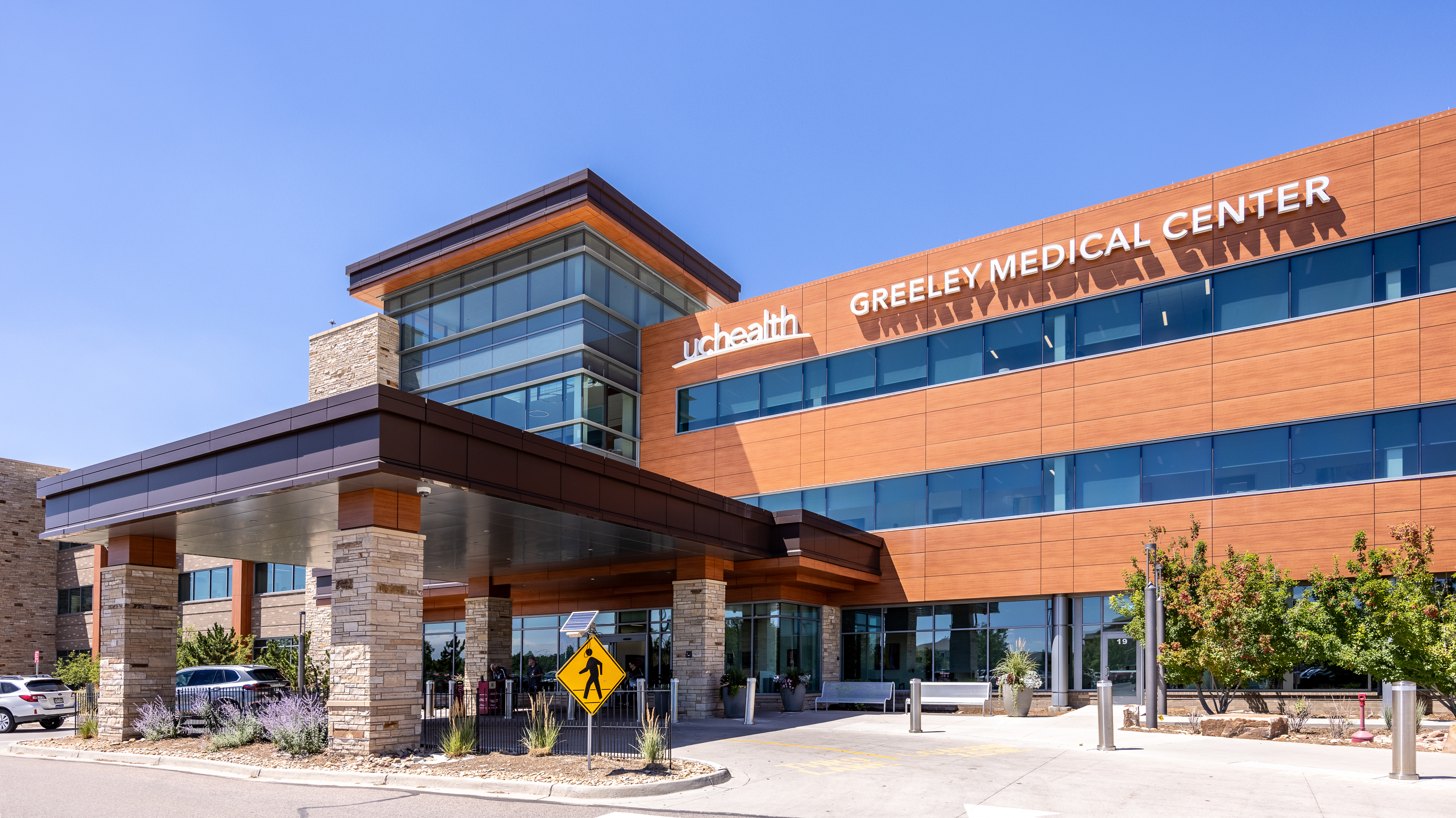 Exterior view of UCHealth Greeley Medical Center with wood-toned panels, glass windows, stone columns, and the main entrance canopy.