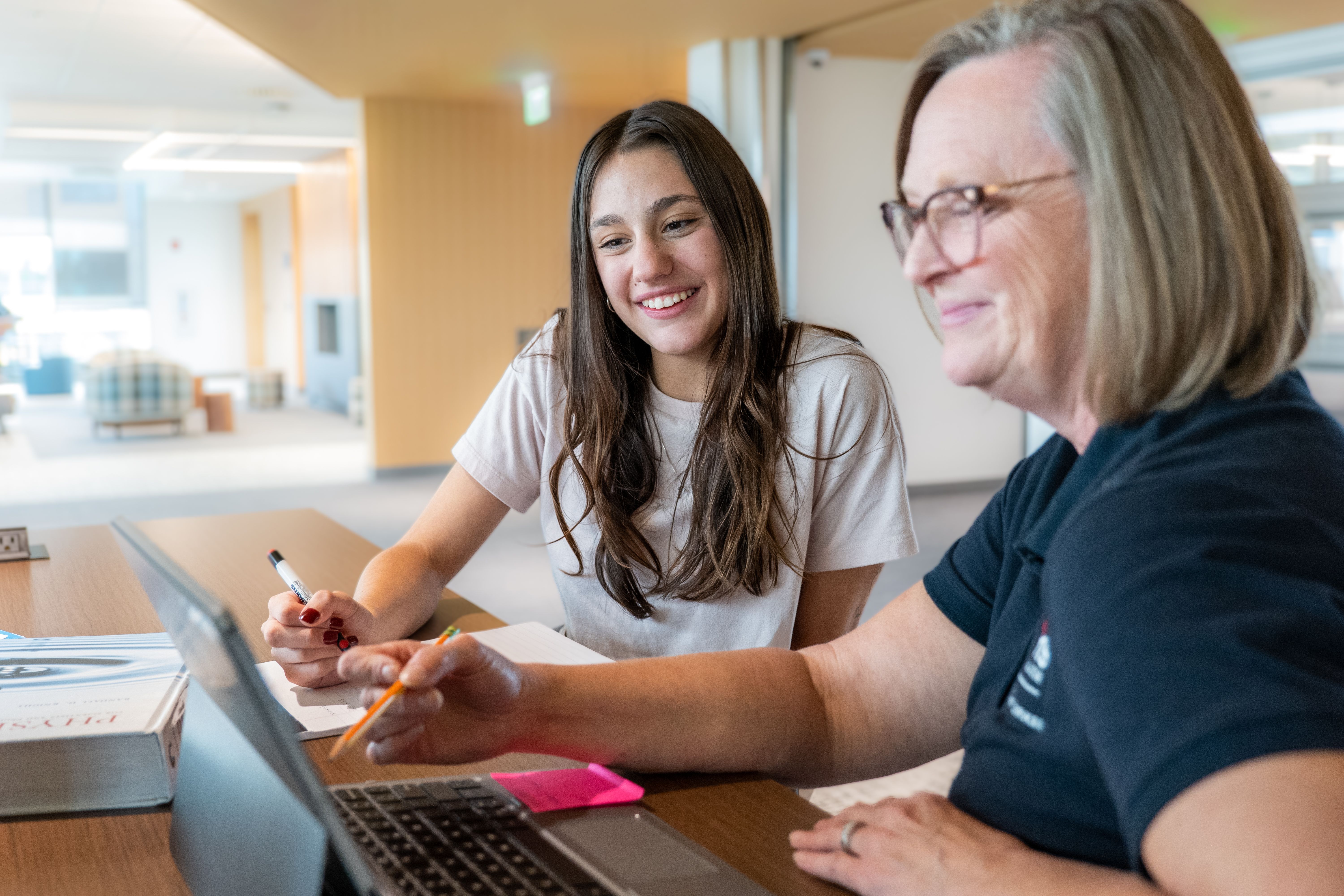 This image depicts a student at Aims Community College receiving support from a tutor.  A student with long, straight brown hair and wearing a white t-shirt is studying the screen of a laptop as the tutor points her attention with a pencil.  The tutor is wearing glasses and a dark polo shirt with an Aims logo on the chest.