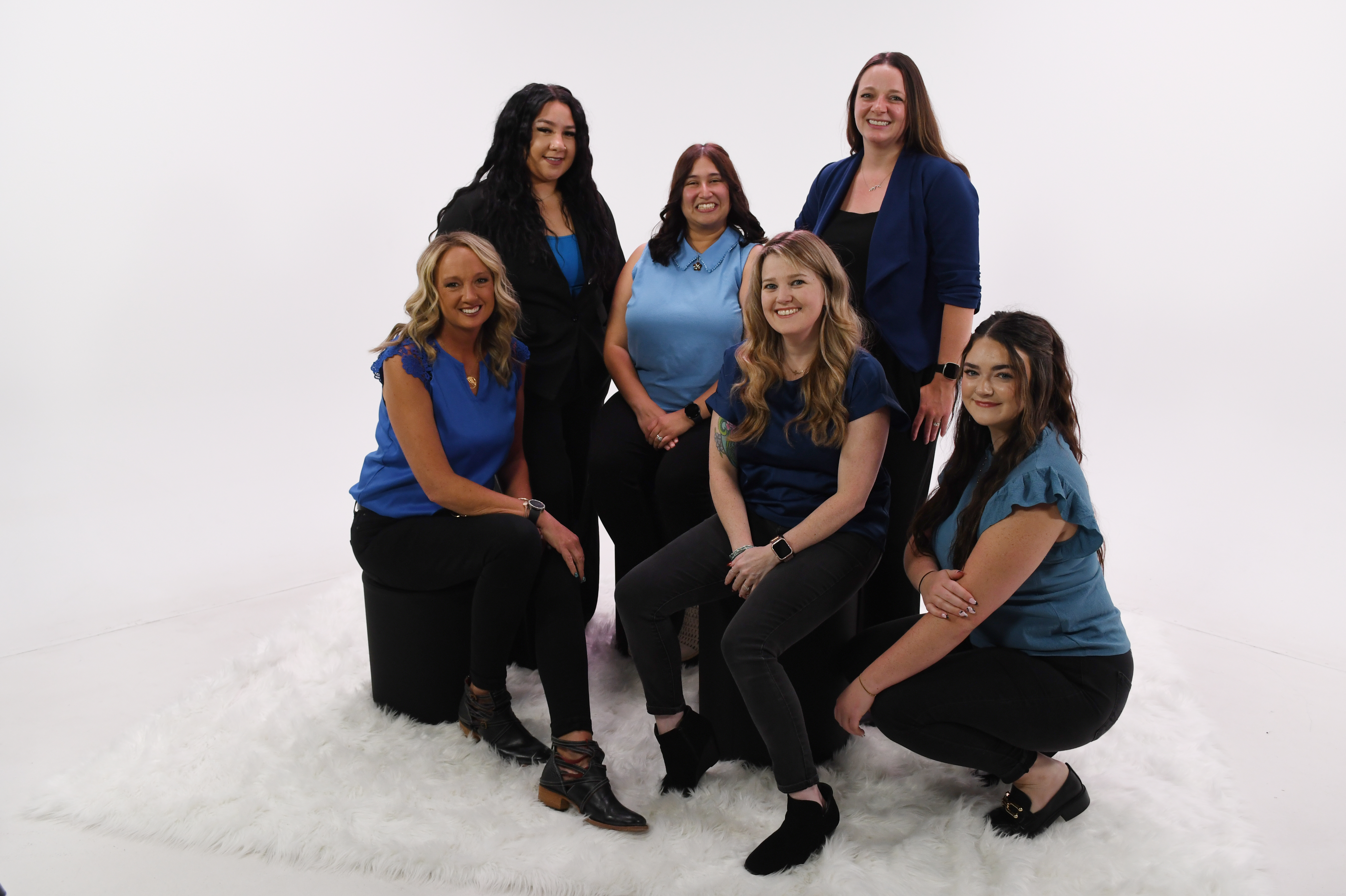 Six women pose in a photo studio with a white background, and white floor. Three of the women are sitting on stools, two are standing and one is crouching. They are all wearing blue shirts and black pants, with one of the standing ladies wearing a black blazer.  