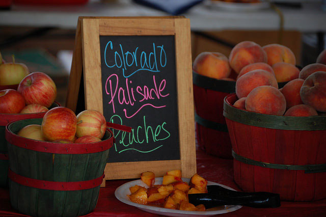 Fresh Palisade peaches and apples displayed in wooden baskets at a farmers market with a chalkboard sign.
