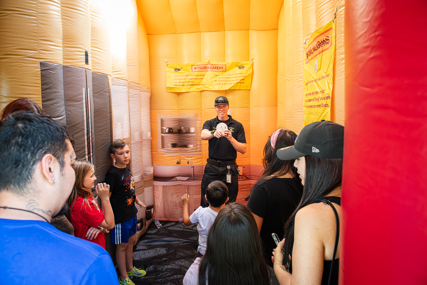 A Greeley Fire staff member holds up a smoke detector and points at a child in the audience of a fire safety presentation held in an inflatable fire safety house. 