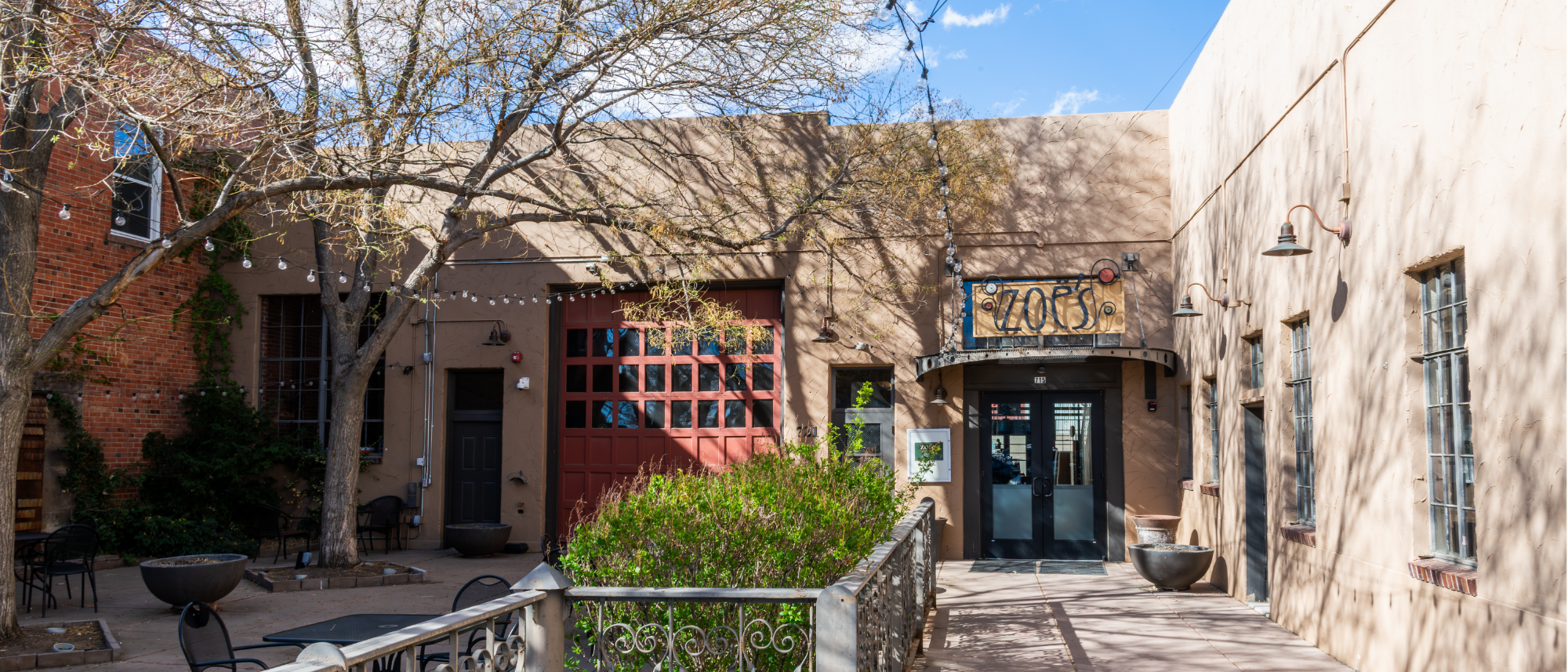 Exterior entrance of Zoe’s Cafe and Events with a courtyard and trees. There is a large sign above the entrance in yellow that says “Zoe’s”. 