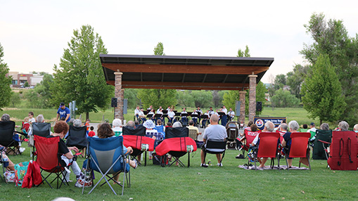 Community members gather on a grassy lawn with folding chairs to enjoy a live outdoor concert performance under a pavilion in a park setting.