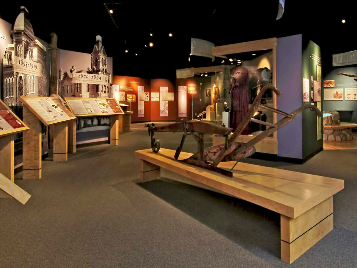 Historical objects and images displayed in a large museum gallery. A large plow stands on a display pedestal in the foreground