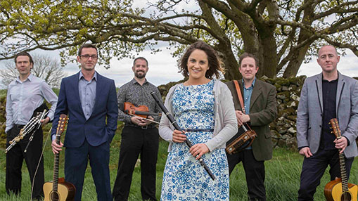 A group of musicians standing outdoors with various instruments, featuring a backdrop of trees and grass.