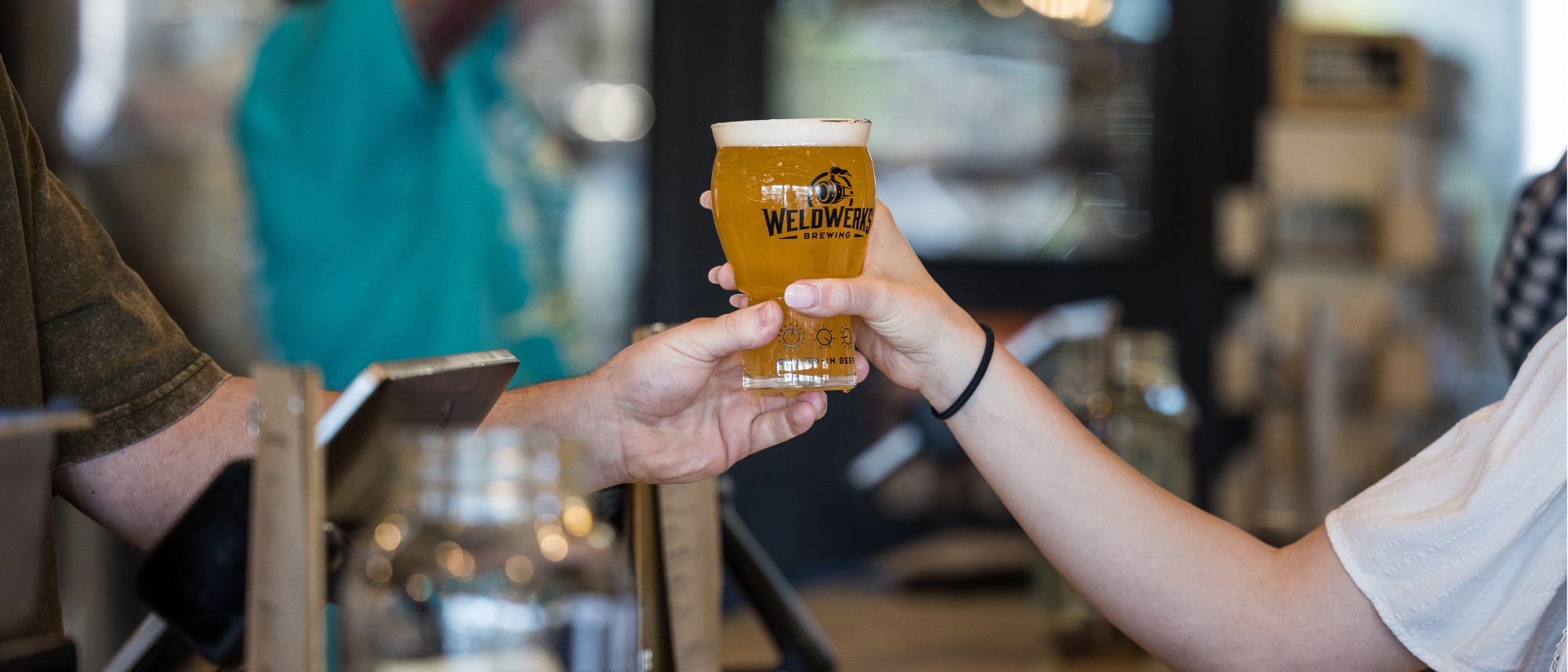WeldWerks Brewing Co brand pint glass full of beer being passed to a customer showing their logo.